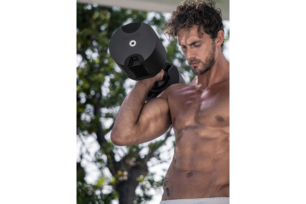 Shirtless man holding black adjustable dumbbell at shoulder height outdoors with trees in background
