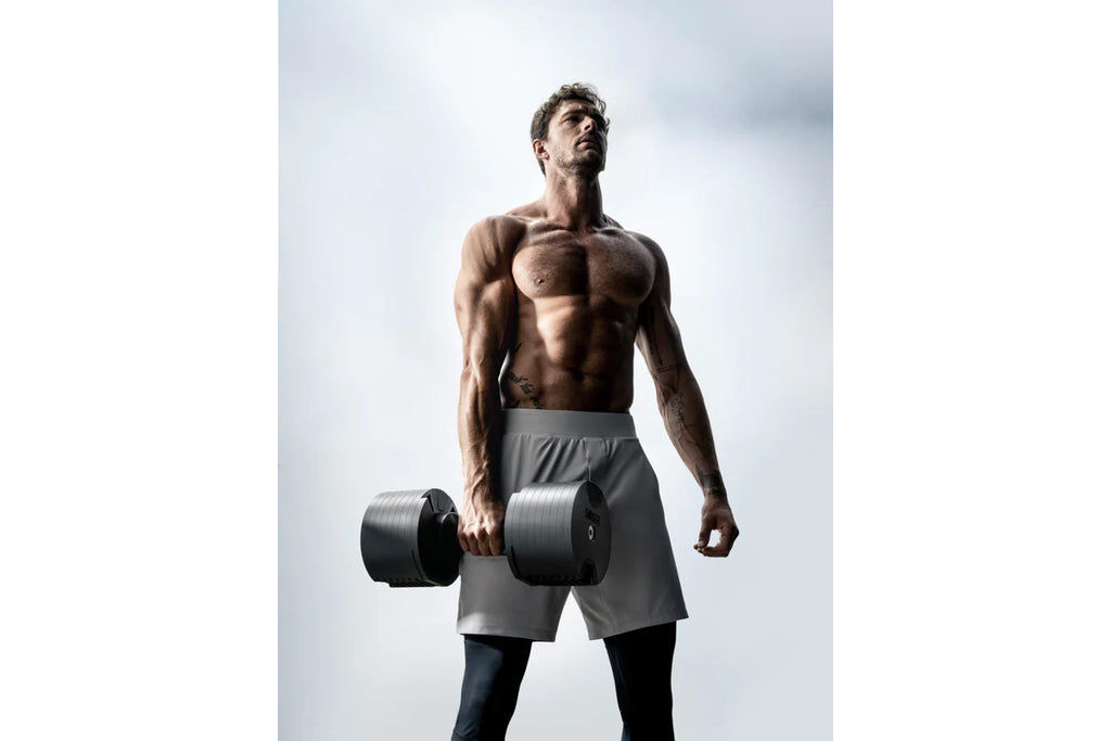 Shirtless man holding black adjustable dumbbells wearing gray shorts against white background