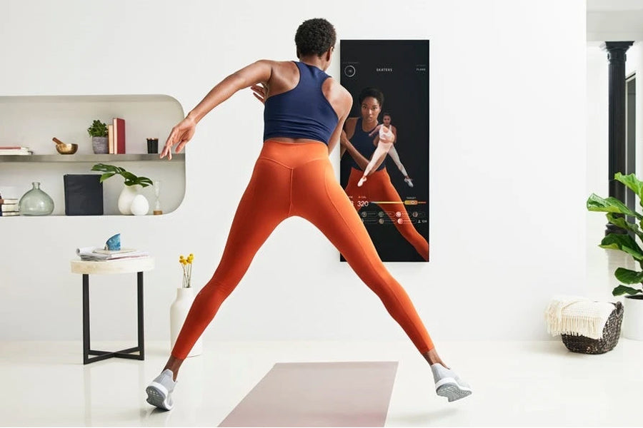Person wearing navy tank top and orange leggings doing a lunge in front of black Studio Mirror displaying workout content on 
