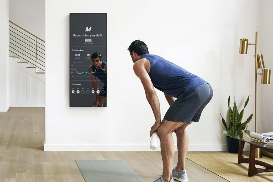 Man in blue tank top and black shorts exercising in front of dark digital fitness mirror mounted on white wall in modern home